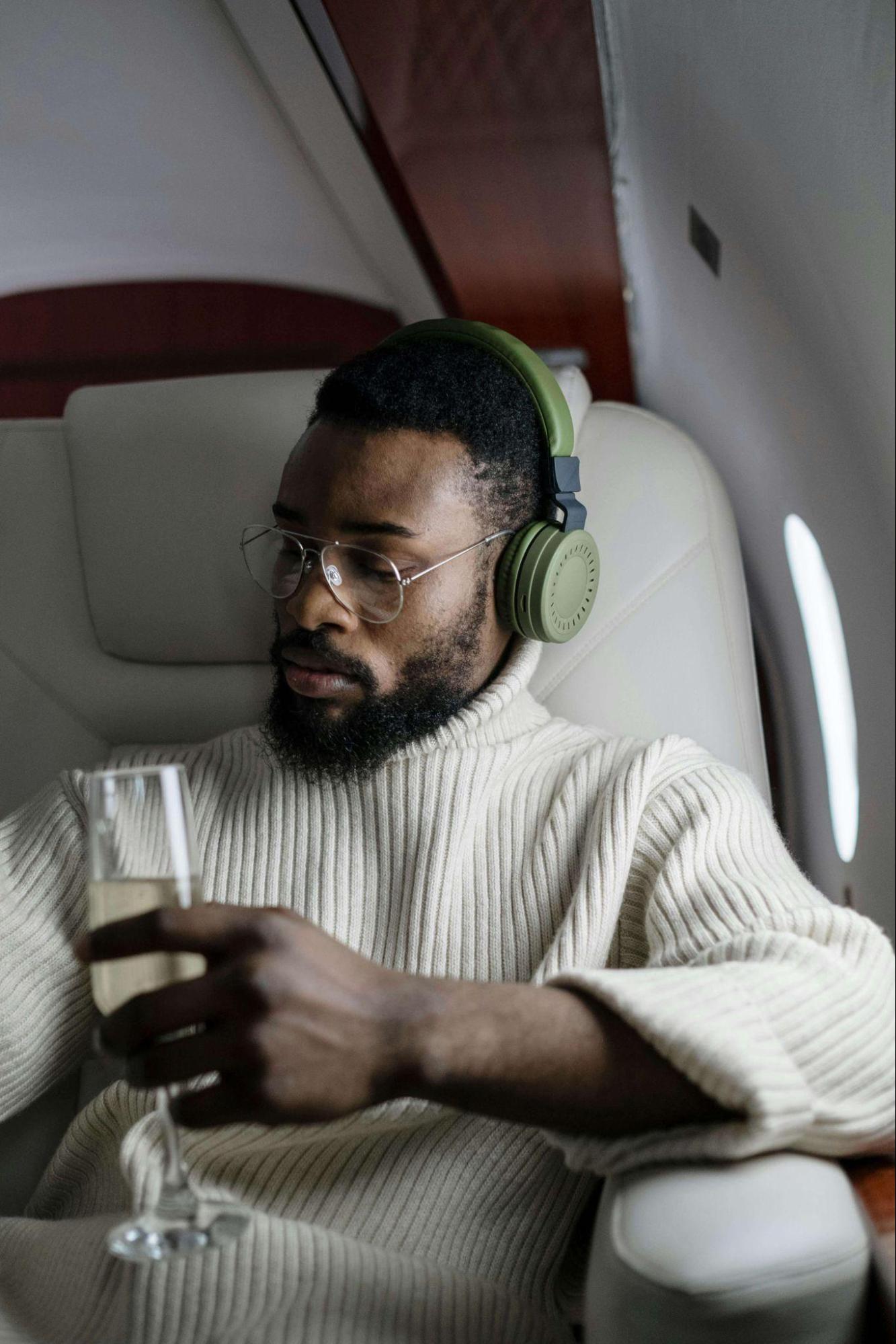 A man in a cream sweater sips champagne and wears headphones on a private flight, relaxed and immersed in the luxury travel experience