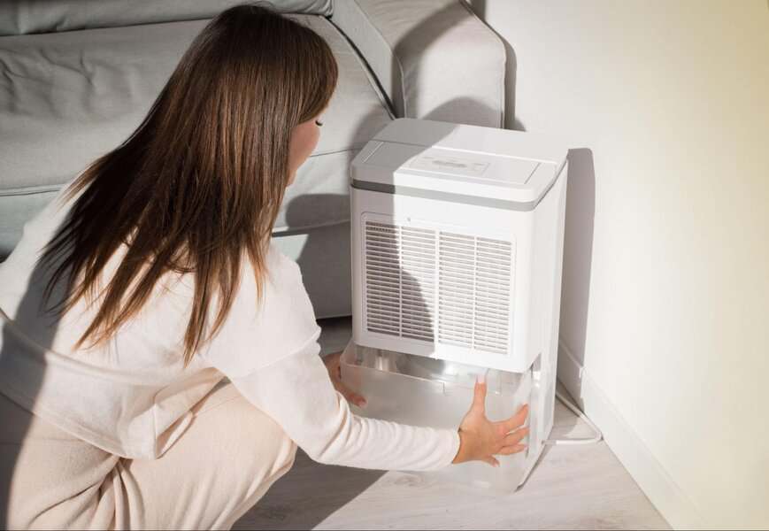 Woman kneels next to a dehumidifier, taking out the water tank in a bright living room to improve air quality