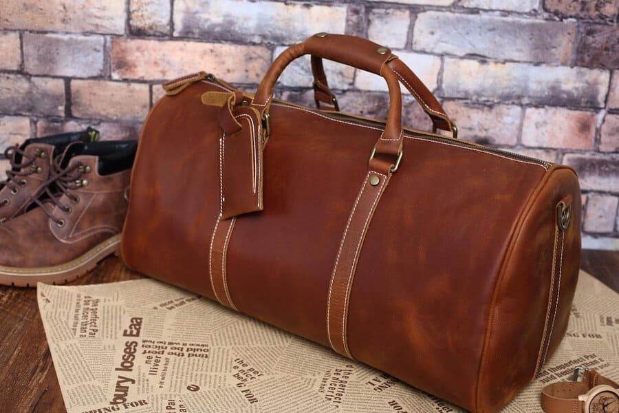 Brown leather duffel bag with handles and tag, ideal for keeping suits smooth, set by boots beside a brick wall