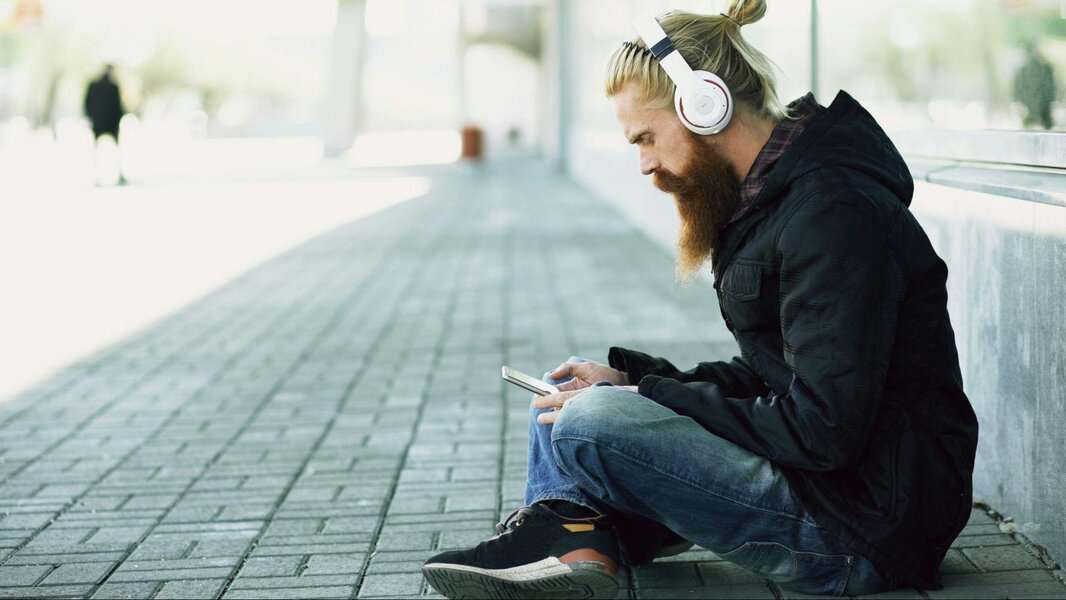 Man with headphones and a beard sits on pavement in luxury, wearing a black jacket and jeans, looking at his phone