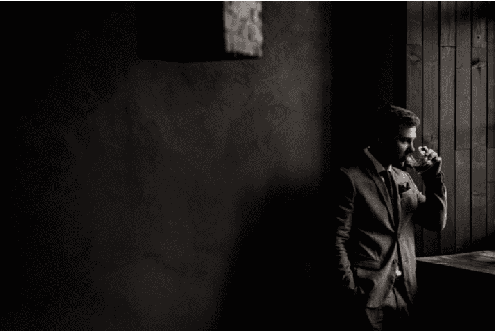 A man in a tuxedo stands by a window drinking from a glass, James Bond style, in a dimly lit room with dark walls