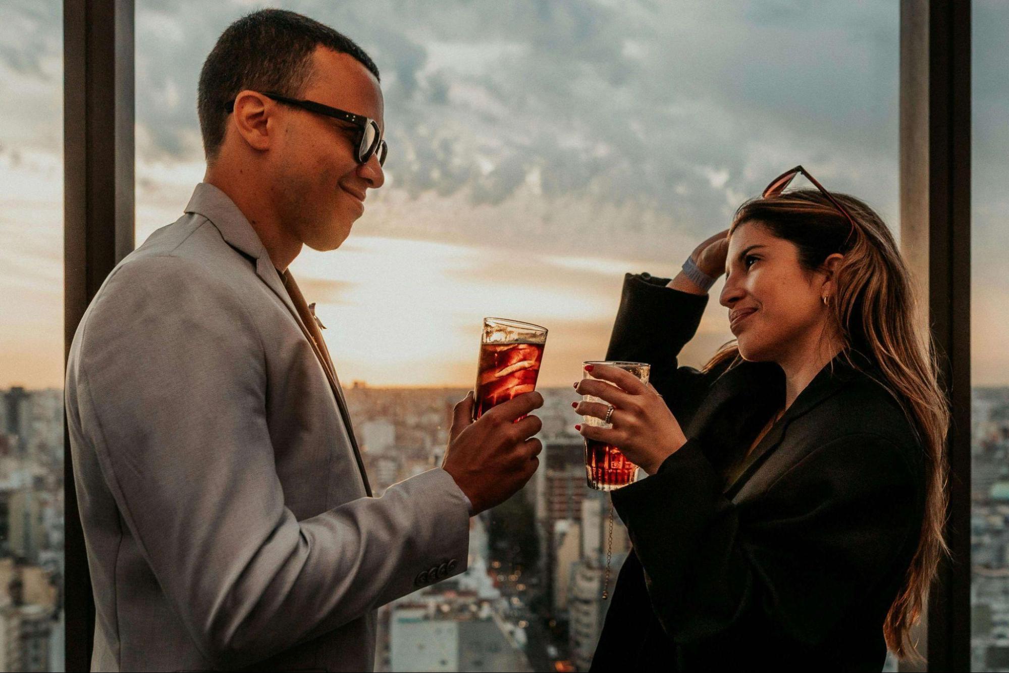 A man and woman clink glasses at sunset on a rooftop, city skyline glowing behind them as they revel in luxurious travel elegance