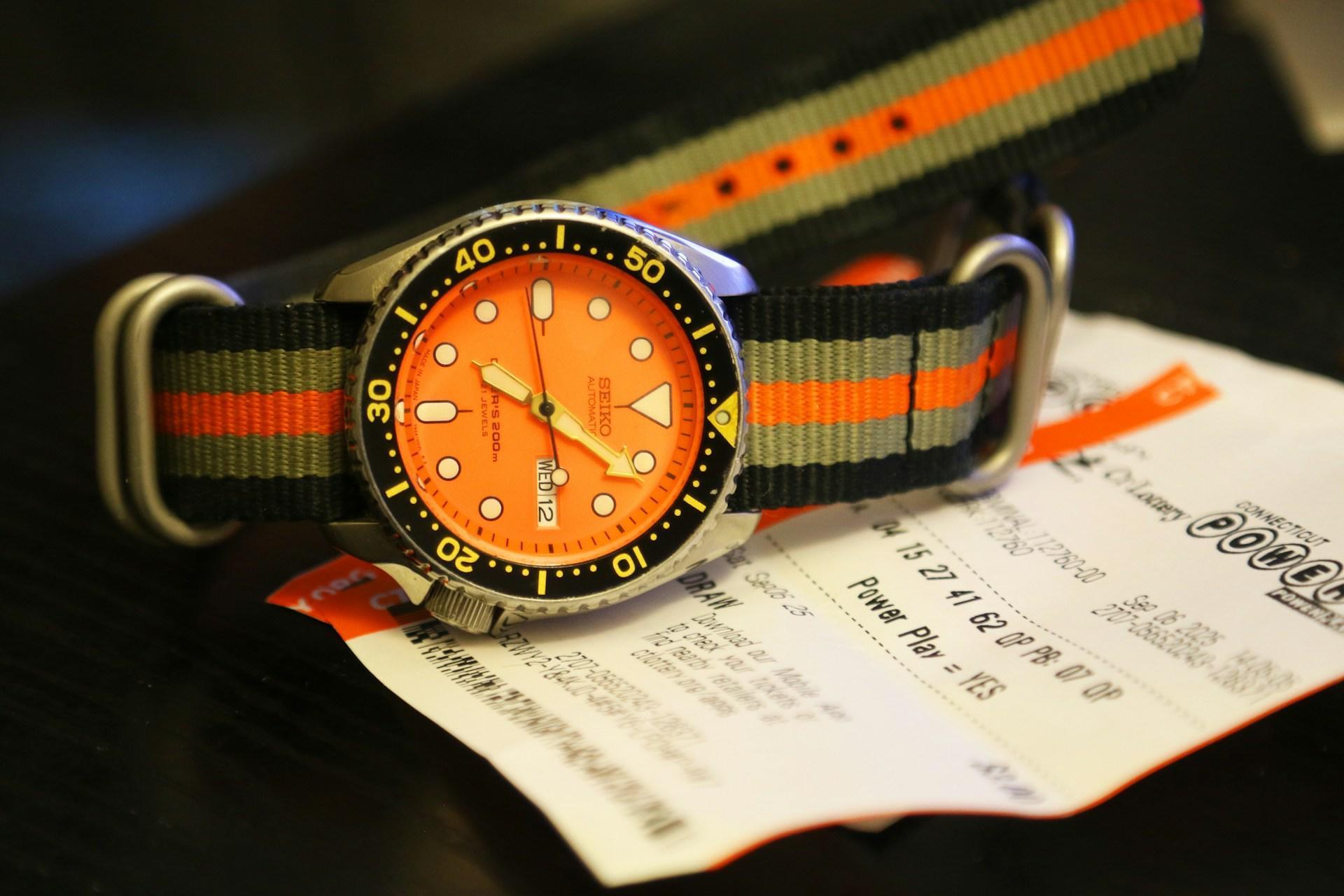 A wristwatch with an orange face and sporty nylon strap lies on a lottery ticket placed on a dark surface