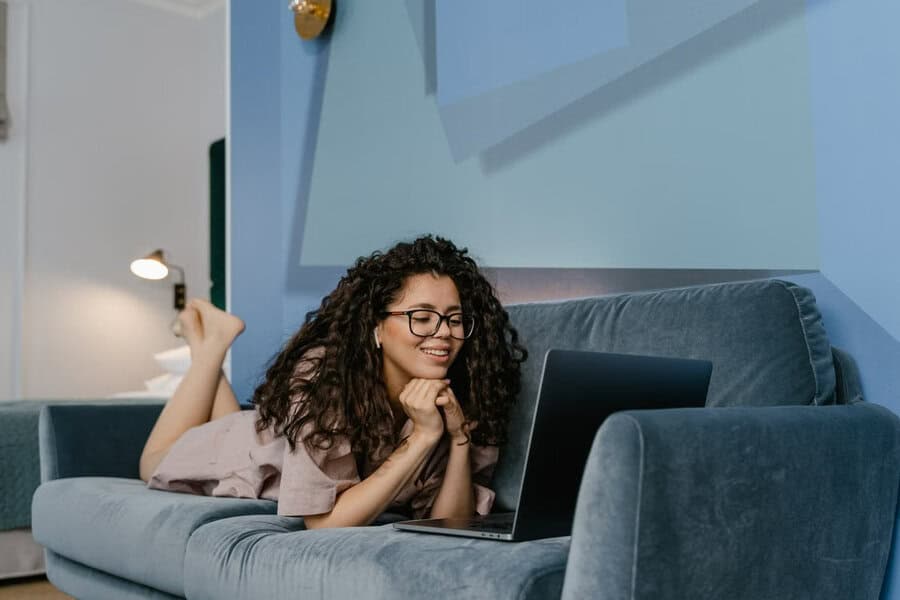 Woman with curly hair and glasses relaxes on a blue couch, smiling at her laptop as she discovers Emerald Chat’s interest-based matching