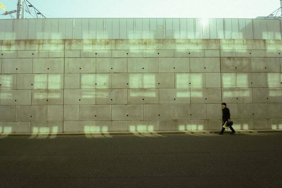 A person in a wrinkle free suit walks along a street next to a tall, windowless concrete wall under pale sunlight