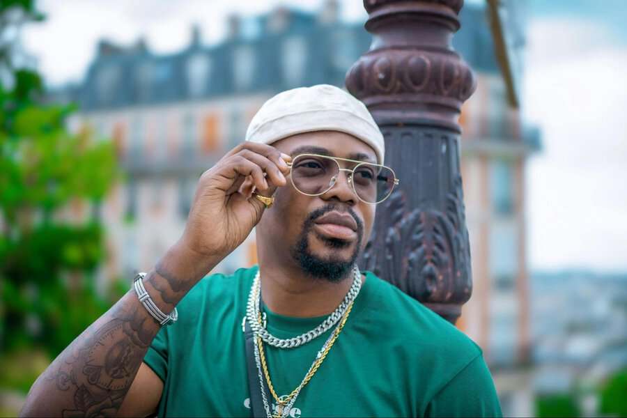 Man in a green shirt and cap adjusts his glasses, wearing sturdy jewelry, outdoors by a lamp post with buildings in the background