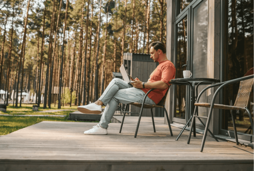 Man using a laptop on a wooden deck, soaking in the outdoors near his cozy cabin and surrounded by nature