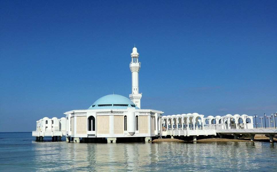 A white mosque with a blue dome stands above the Red Sea, connected by an arched walkway—a highlight on any Jeddah trip