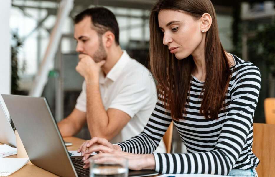 Two people work at a table; she types on her laptop while he looks at a Thisromance review on his computer screen