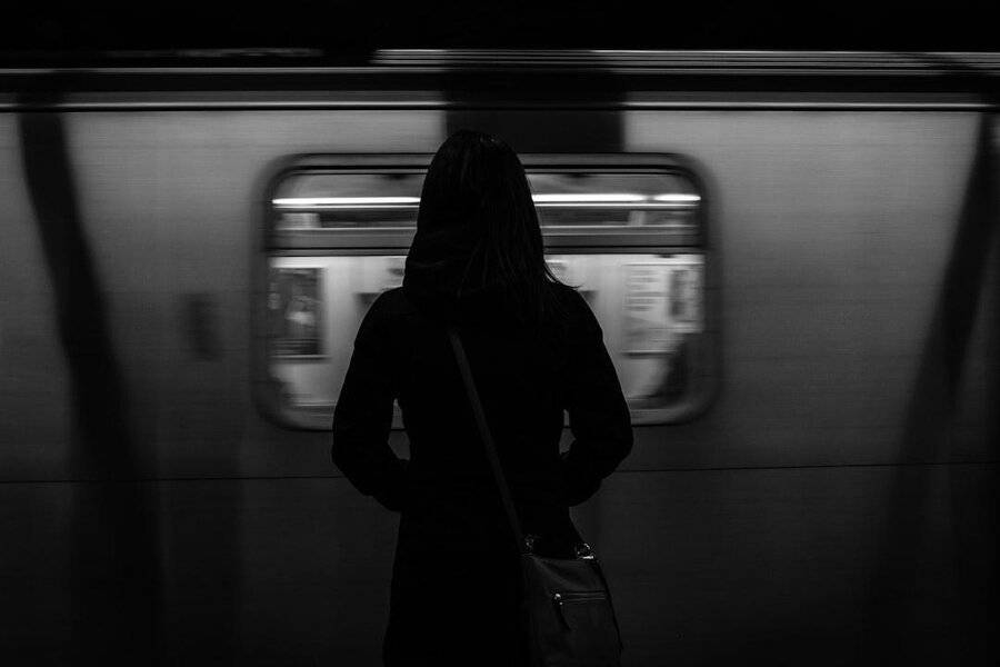 A person in a suit waits on a dim subway platform, looking toward a passing train with blurred windows, capturing the city commute