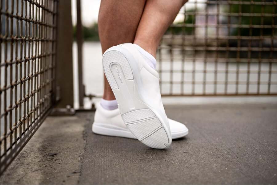 Person wearing Amberjack Low Top leather sneakers stands on concrete near a metal fence, legs crossed, water visible in the background