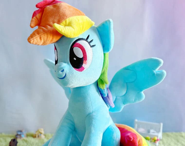 A Rainbow Dash plush toy with rainbow hair and a matching hat sits on a colorful surface, perfect for fun room décor