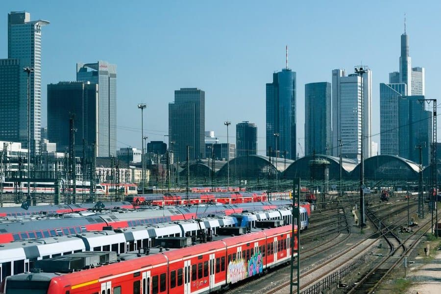 Red trains at a busy station, skyscrapers behind—a great commute spot for suits on a sunny day under clear blue skies