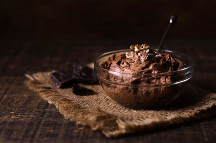 Glass bowl of chocolate mousse with chopped nuts for holiday treats sits on burlap mat, surrounded by pieces of chocolate
