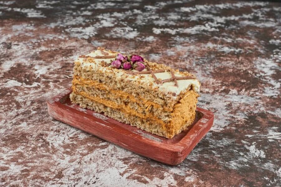 Slice of layered cake with cream and nuts, topped with pink flowers on a tray, set against a marbled background