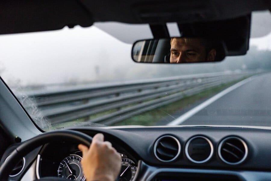 Inside a car, a commuter in a suit drives along a foggy road; their face is visible in the rearview mirror