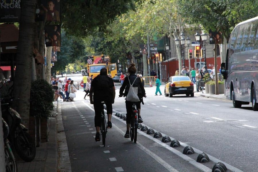 Two people in suits ride bikes down a city street lined with leafy trees, passing cars, pedestrians, and buses