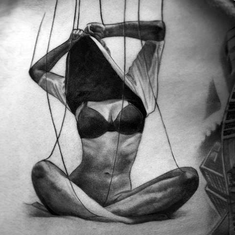 Realistic puppet tattoo of a woman sitting cross-legged in lingerie, controlled by strings, symbolizing vulnerability or constraint.