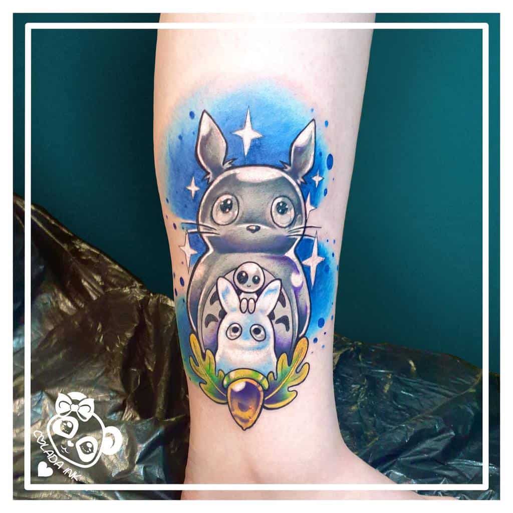 Colorful Totoro leg tattoo with a cosmic blue background, stars, and Totoro family layers, accented by an acorn and leaves