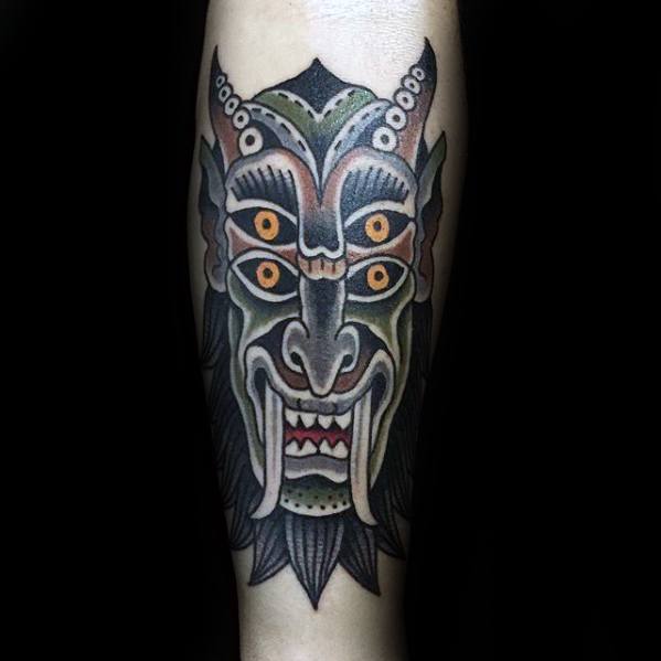 Traditional Krampus tattoo featuring a symmetrical horned face with six orange eyes, sharp fangs, and bold black and green shading.