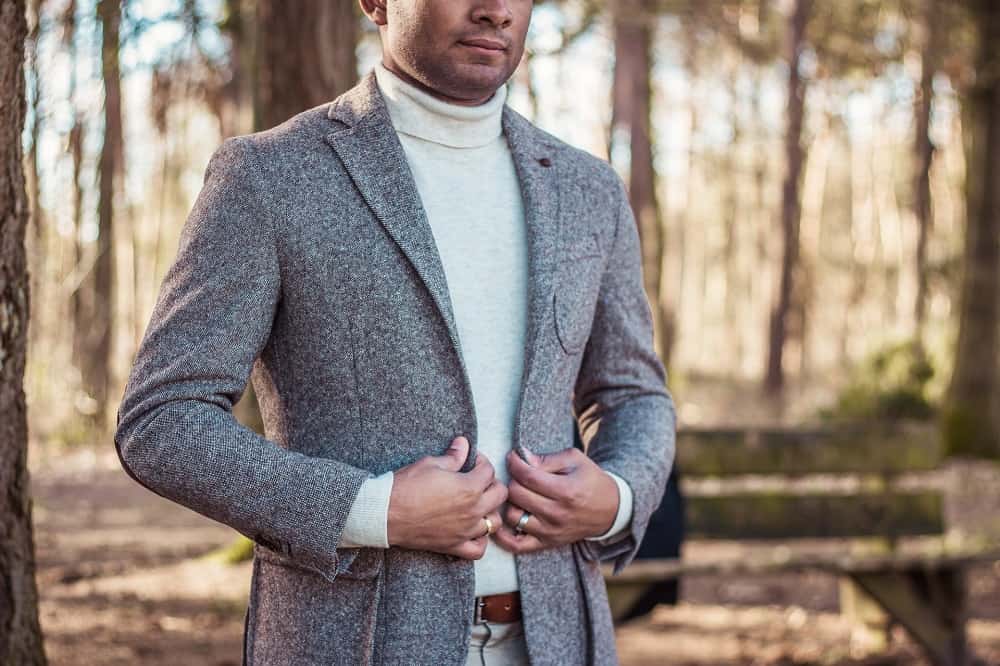 man standing in the forest poses, wearing a grey trench coat and white turtleneck