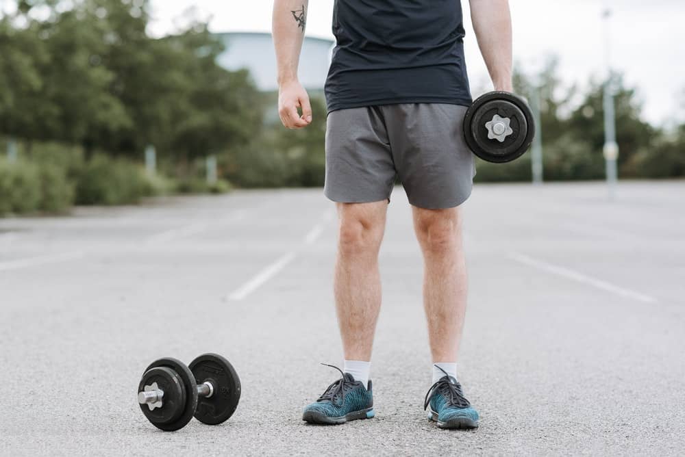 man stands alone in an empty parking lot, one dumbbell by his side in hand, the other on the ground by his feet