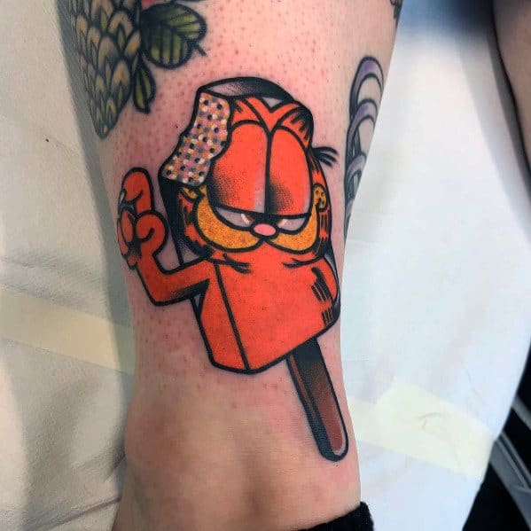 Tattoo of an orange popsicle designed like a cartoon cat with a playful smirk, bold black outlines, and a melting ice cream topping.