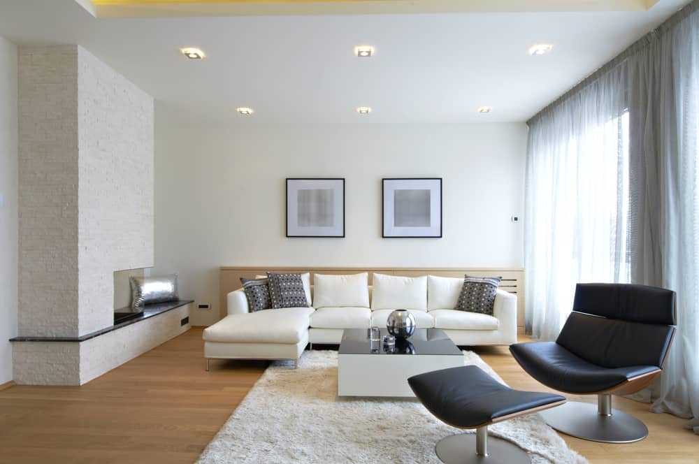 Modern living room with a white sectional sofa, black leather chairs, stone fireplace, and soft natural lighting