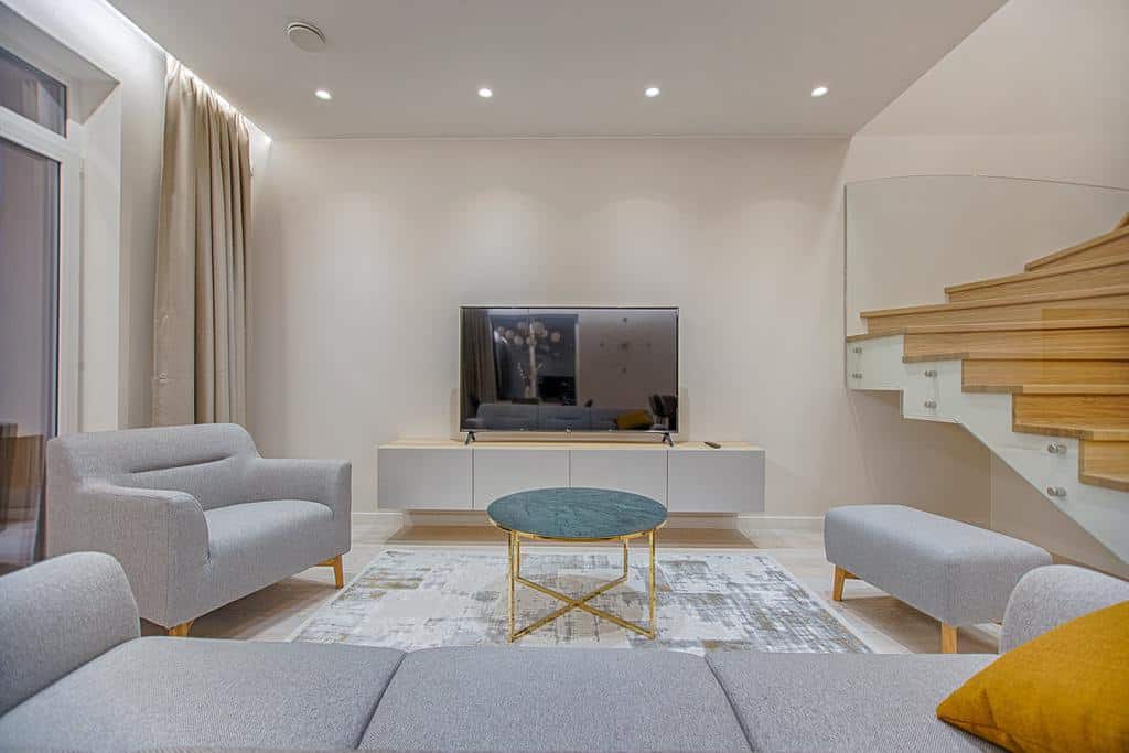 Minimalist living room featuring soft gray furniture, a flat-screen TV, and a sleek wooden staircase with glass railings