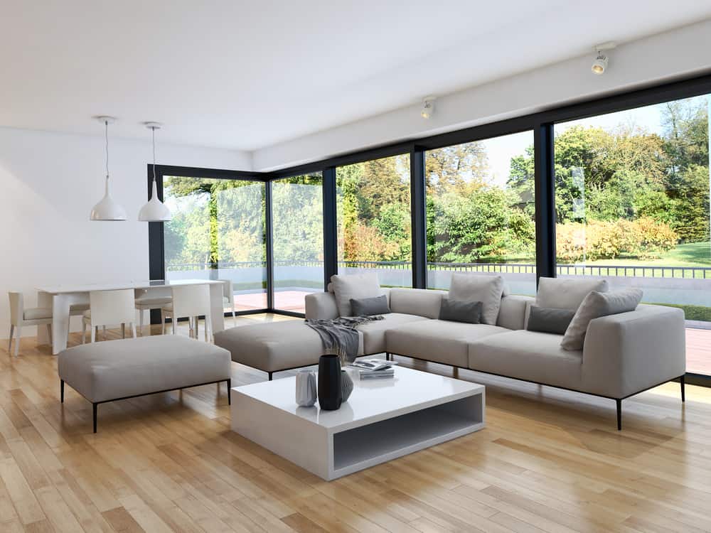 Bright and spacious living room with a neutral minimalist design, featuring a large cream sectional sofa, light wooden floors, and floor-to-ceiling windows offering a view of a lush outdoor garden