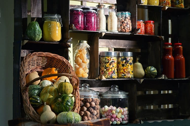 Wooden shelves with jars of dried goods, preserves, and basket of gourds in rustic pantry.