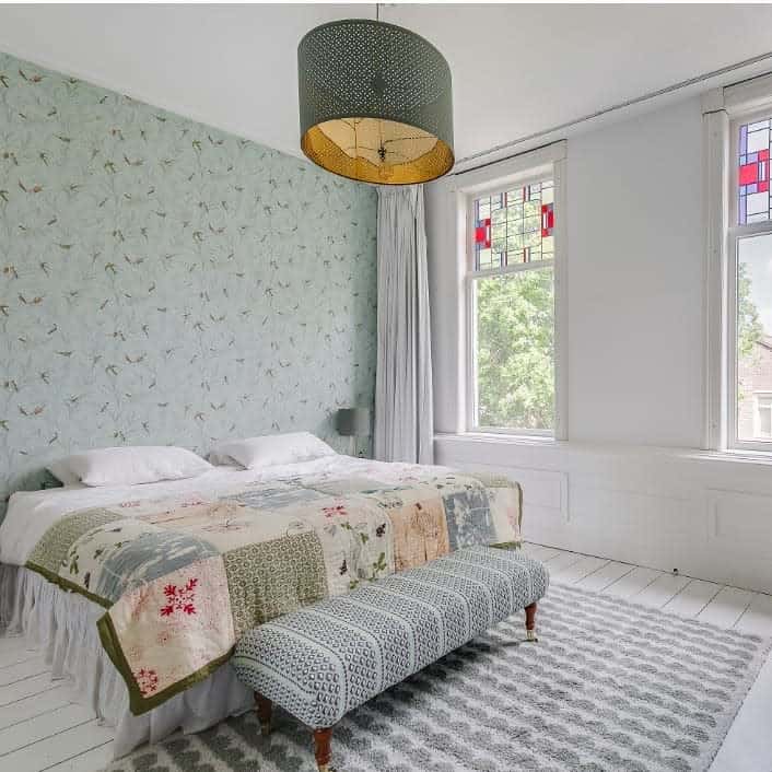 Country-style bedroom with green bird wallpaper, patchwork quilt, and patterned ottoman.