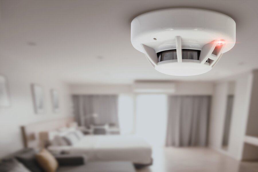 Close-up of a smoke detector with a red light on, mounted on the ceiling of a modern, blurred bedroom