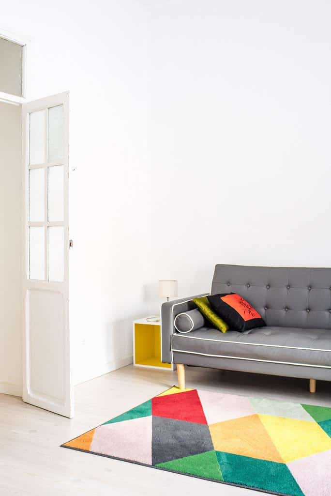 Bright minimalist living room with a gray tufted sofa, colorful geometric rug, and yellow side table for a playful touch