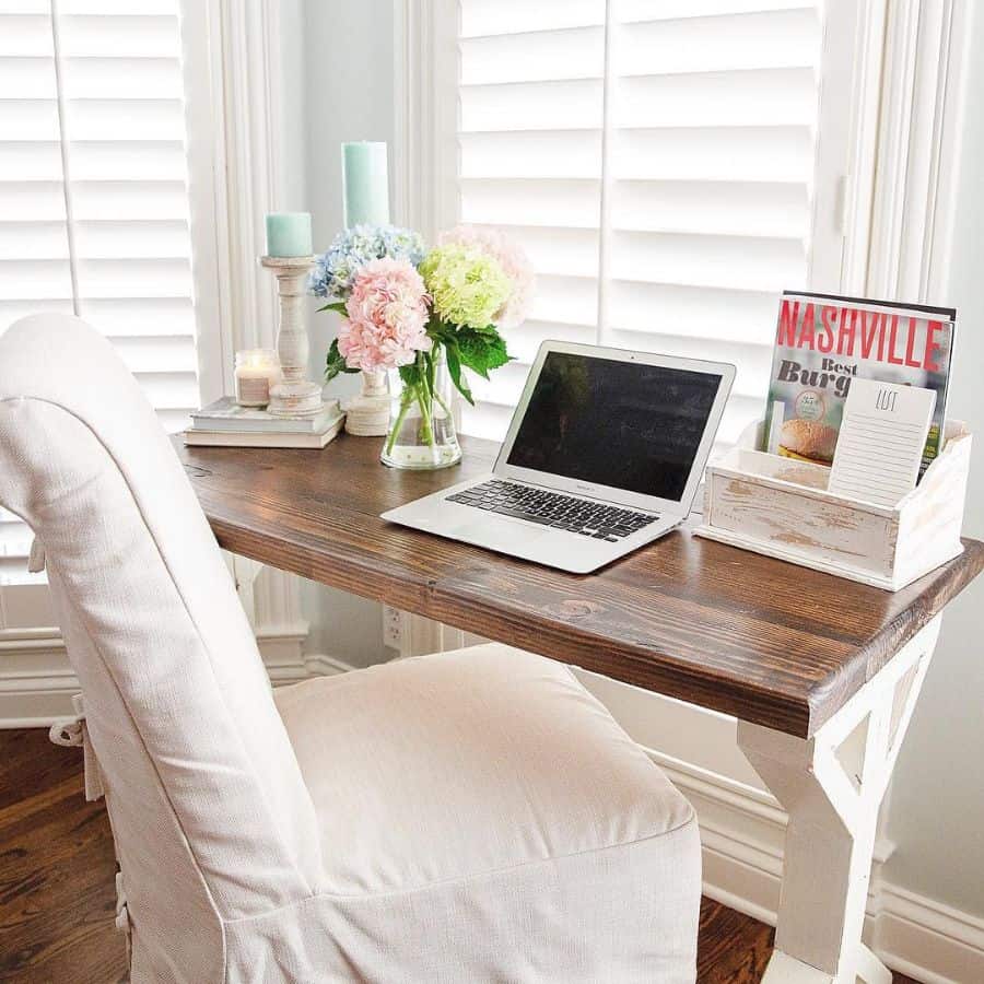 Rustic wood desk with laptop, flowers, and magazines in a bright bedroom with white chair.